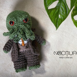Pode incluir: Um brinquedo de pel&uacute;cia Cthulhu verde feito de croch&ecirc; vestindo um terno marrom e uma camisa branca com uma gravata laranja. O brinquedo tem olhos vermelhos e tent&aacute;culos. O brinquedo est&aacute; sentado em uma superf&iacute;cie bege com uma planta verde no fundo. O texto "Noctua Art & Crafts" &eacute; vis&iacute;vel no canto inferior direito da imagem.