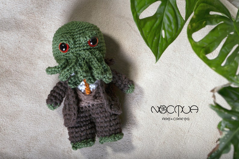 Pode incluir: Um brinquedo de pel&uacute;cia Cthulhu verde feito de croch&ecirc; vestindo um terno marrom e uma camisa branca com uma gravata laranja. O brinquedo tem olhos vermelhos e tent&aacute;culos. O brinquedo est&aacute; sentado em uma superf&iacute;cie bege com uma planta verde no fundo. O texto "Noctua Art & Crafts" &eacute; vis&iacute;vel no canto inferior direito da imagem.