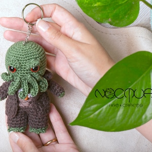 Pode incluir: Chaveiro Cthulhu de croch&ecirc; verde com olhos vermelhos e um terno de croch&ecirc; marrom. O chaveiro &eacute; segurado na m&atilde;o e tem um chaveiro prateado.