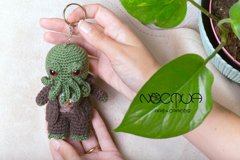 Pode incluir: Chaveiro Cthulhu de croch&ecirc; verde com olhos vermelhos e um terno de croch&ecirc; marrom. O chaveiro &eacute; segurado na m&atilde;o e tem um chaveiro prateado.