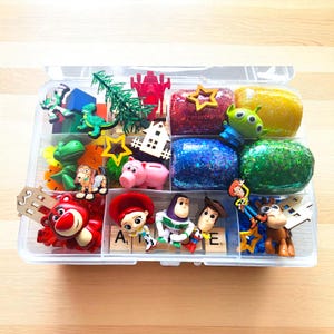 May include: A clear plastic organizer filled with colorful character figurines and glittery, textured shapes. The collection includes Toy Story characters, a green dinosaur, a pink pig, and star-shaped accents. The letters "A" and "E" are visible.