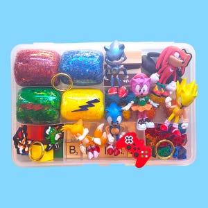 Puede incluir: Un organizador de pl&aacute;stico transparente lleno de art&iacute;culos coloridos con el tema de Sonic the Hedgehog. Incluye figuras de personajes, bloques brillantes, anillos y un mando de juego rojo. El fondo es azul claro liso.