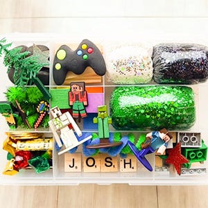 May include: A clear plastic storage box filled with various items, including green and black play dough, glitter, a green tree, a green and white Minecraft character, a green and brown zombie character, a red and brown Minecraft character, a green and brown video game controller, and a set of letter tiles spelling "JOSH".