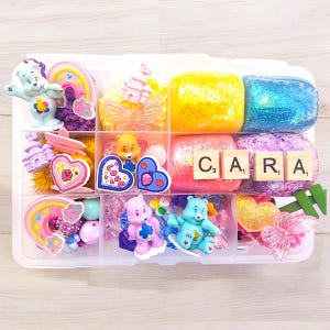 May include: A clear plastic organizer filled with colorful craft supplies. The compartments hold Care Bear figurines, heart-shaped decorations, glittery slime, and wooden letter tiles spelling "CARA". The overall theme is a collection of cute, colorful items.
