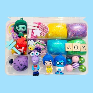 May include: A clear plastic organizer filled with colorful charms and decorative elements. Includes character figurines, glittery blocks, and wooden letter tiles spelling "JOY". Other items include a star, a bee, and the text "Don't trip over what's behind you."