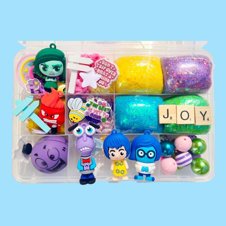 May include: A clear plastic organizer filled with colorful charms and decorative elements. Includes character figurines, glittery blocks, and wooden letter tiles spelling "JOY". Other items include a star, a bee, and the text "Don't trip over what's behind you."