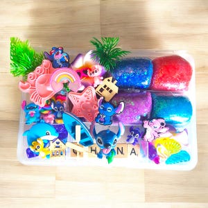 May include: A clear plastic container filled with colorful craft supplies. The container holds various character-themed charms, including Stitch, a rainbow, and a starfish. Also included are several containers of glittery slime in blue, red, purple, and teal.