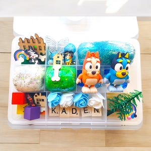 May include: A clear plastic storage box filled with various play items, including blue and green play dough, a small wooden house, a blue and white butterfly, a rainbow, a wooden fence, a small dog figurine, a blue and white glitter slime, a blue and yellow dog figurine, a red and orange dog figurine, a small green tree, white and blue flowers, and letter tiles spelling "KADEN".