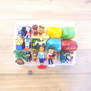 May include: A clear plastic container filled with colorful play dough and various toy figurines. The figurines include cartoon characters, vehicles, and small houses. The play dough is in yellow, green, blue, and red.