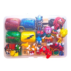 Puede incluir: Un recipiente de pl&aacute;stico transparente lleno de varios art&iacute;culos tem&aacute;ticos de Sonic the Hedgehog. Incluye figuras de personajes de Sonic, Tails, Knuckles y Amy Rose, junto con bloques brillantes, anillos y un mando de juego rojo. El recipiente tambi&eacute;n tiene fichas de Scrabble.
