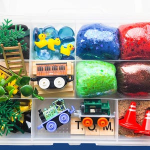 May include: A clear plastic storage container with multiple compartments filled with various toys and playthings. The compartments contain green and yellow plastic trees, a wooden train, a blue toy train, yellow rubber duckies, blue and green slime, brown sand, and red and white traffic cones.