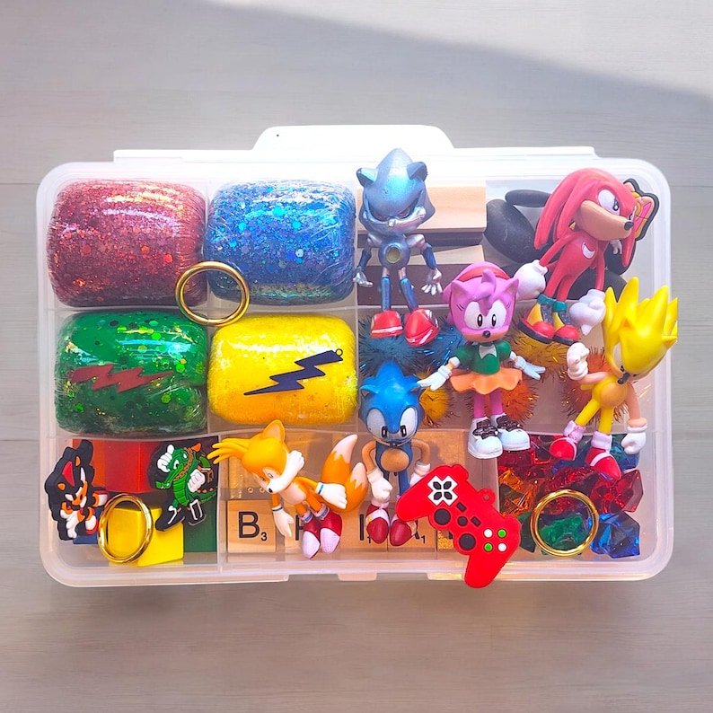 Puede incluir: Un organizador de pl&aacute;stico transparente lleno de figuras de Sonic the Hedgehog y bloques coloridos y brillantes. La colecci&oacute;n incluye a Sonic, Tails, Knuckles, Amy Rose y Super Sonic, junto con un mando de juego rojo y anillos dorados. Los bloques son rojos, azules, verdes y amarillos.