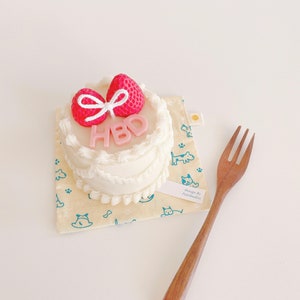 May include: A small white cake with pink frosting and a pink bow on top. The cake is decorated with the letters "HBD" and is sitting on a yellow and blue patterned surface. The cake is designed by PetStudios.