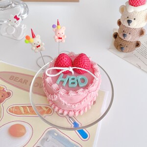 May include: A pink frosted cake with white frosting and two red strawberries on top. The cake is decorated with a white ribbon and the letters "HBD" in light blue frosting. The cake is on a clear glass cake stand.