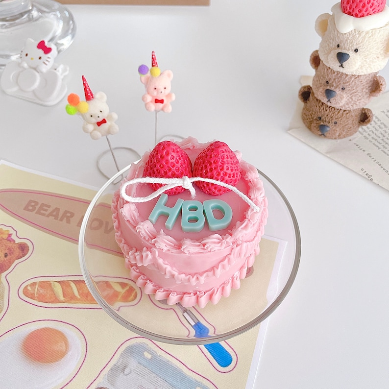 May include: A pink frosted cake with white frosting and two red strawberries on top. The cake is decorated with a white ribbon and the letters "HBD" in light blue frosting. The cake is on a clear glass cake stand.