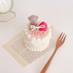 May include: A small white cake with pink frosting and a teddy bear figurine on top. The cake is decorated with pink letters that spell out "HBD".