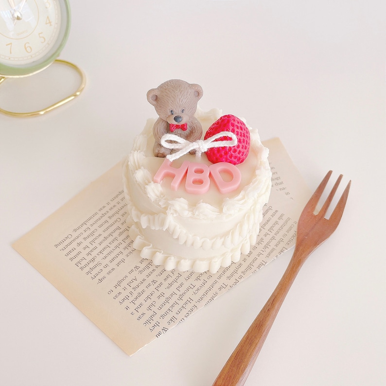 May include: A small white cake with pink frosting and a teddy bear figurine on top. The cake is decorated with pink letters that spell out "HBD".
