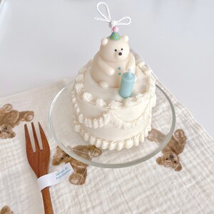 May include: A white cake decorated with white icing and a white polar bear figurine wearing a party hat. The cake is on a clear glass plate and a white cloth background.