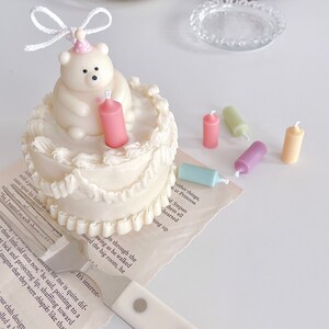 May include: A white cake with white frosting and a white polar bear figurine on top. The cake has a pink candle in the centre and is surrounded by colourful candles on a white surface.