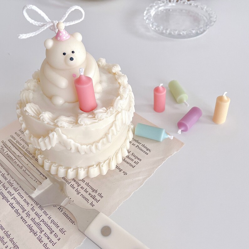 May include: A white cake with white frosting and a white polar bear figurine on top. The cake has a pink candle in the centre and is surrounded by colourful candles on a white surface.