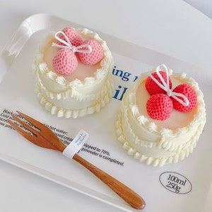 May include: Two small white cakes decorated with white frosting and pink strawberry accents. The cakes are on a white plate with a wooden fork. The text "Design By Pelo Studios" and "Whipped to 70% powder is completed." is on a white label. The text "100ml 250g" is on a white label.