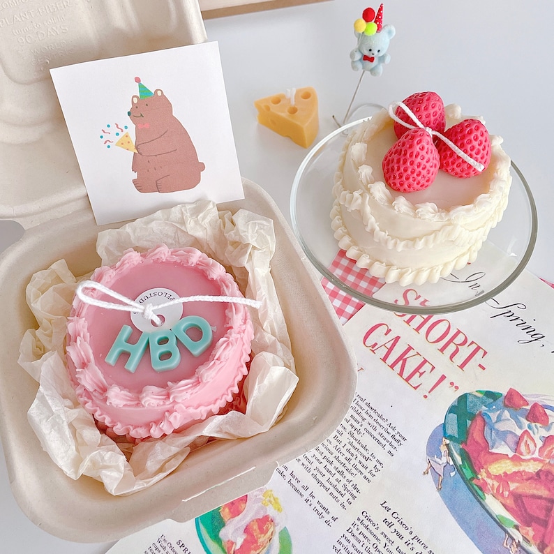 May include: Two pink and white birthday cake candles with a blue and green "HBO" inscription. One candle is in a white box with a white and pink paper liner. The other candle is on a white plate with a pink and white checkered pattern. The background includes a book with the text "In Spring. Short-Cake!" and a small bear figurine.