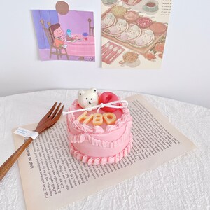 May include: A small pink cake with white frosting and the letters "HBD" in gold. A small white bear figurine and a red apple are on top of the cake.