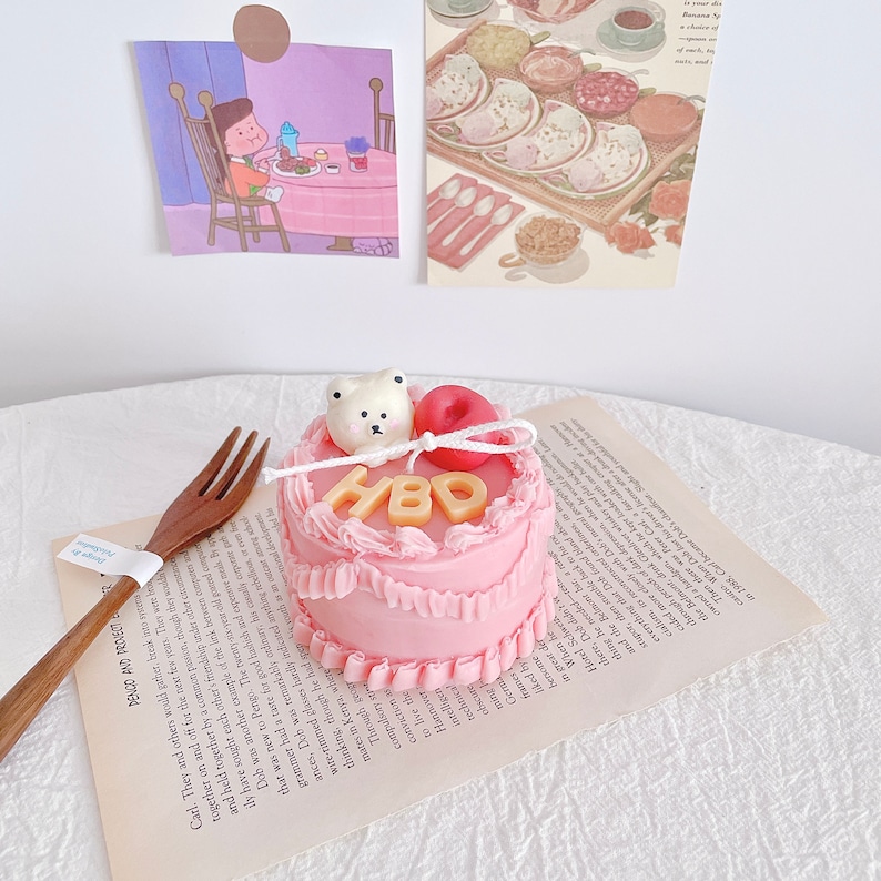 May include: A small pink cake with white frosting and the letters "HBD" in gold. A small white bear figurine and a red apple are on top of the cake.