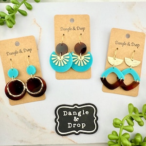 May include: Three pairs of earrings on display. Each pair is attached to a brown card with the text "Dangle & Drop". The earrings feature gold-colored hooks and accents, with turquoise, brown, and gold-colored geometric designs.