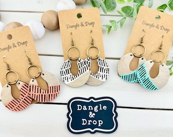 May include: Wooden dangle earrings with brass-colored hoops. The earrings feature a circular design with painted sections in coral, black and white, and turquoise. The earrings are displayed on brown tags with the text "Dangle & Drop".