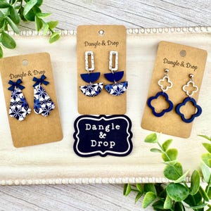 Puede incluir: Tres pares de pendientes en exhibición. Uno presenta un diseño de lazo, otro tiene un patrón de semicírculo y floral, y el último tiene forma de trébol. Todos los pendientes son azules y blancos, con herrajes plateados. La marca "Dangle & Drop" es visible.
