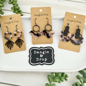 Peut inclure: Trois paires de boucles d'oreilles présentées sur des cartons bruns avec l'inscription "Dangle & Drop". Les boucles d'oreilles présentent des motifs noirs et écaille de tortue dans diverses formes géométriques, notamment des cercles, des carrés et des motifs en forme d'éventail.
