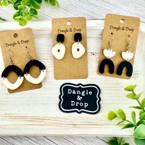 May include: Three pairs of earrings with black and cream-colored designs are displayed on brown cards. The earrings have various shapes, including hoops, abstract shapes, and floral designs. The brand name "Dangle & Drop" is printed on the cards and a black and white sign.