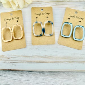 May include: Three pairs of rectangular dangle earrings displayed on brown cardboard cards labelled Dangle & Drop. The first pair is beige with a wood-grain pattern, the second is white with blue spots, and the third is a solid light blue colour.