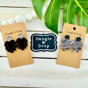 Op de afbeelding: Twee paar bungelende oorbellen op bruine kartonnen displaykaarten met het label Dangle & Drop. Eén paar heeft effen zwarte monstera-bladvormen, terwijl het andere paar een zwart-wit palmbladpatroon heeft. Een centraal bordje vermeldt Dangle & Drop.