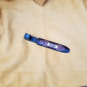 May include: A handcrafted wooden pipe with a dark blue bowl and a decorative, inlaid design on the stem. The stem features a geometric pattern with red, white, and green accents. The pipe rests on a tan leather surface.