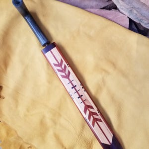 Puede incluir: Un abrecartas de madera hecho a mano con un mango oscuro y un diseño incrustado decorativo. El abrecartas presenta una combinación de madera clara y oscura, con un patrón de flecha geométrica en tonos rojos y marrones. El mango es de color gris oscuro.