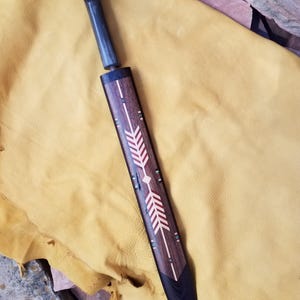Puede incluir: Un cuchillo artesanal con mango oscuro y funda de madera. La funda está decorada con un diseño de flecha en rojo, blanco y verde. El cuchillo y la funda se exhiben sobre una superficie de cuero amarillo.