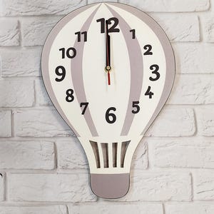 May include: A white and grey hot air balloon clock with black numbers. The clock is shaped like a hot air balloon with a grey top and a white bottom. The numbers are in a circle around the clock face. The clock hands are black.