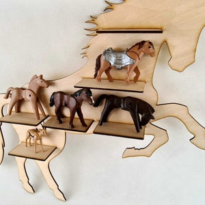 Puede incluir: Estante de madera con forma de caballo con cinco estantes para exhibir caballos de juguete. Los estantes son de madera y están pintados de marrón. El caballo está pintado de marrón y tiene una crin y una cola blancas. Hay cinco caballos de juguete en los estantes, incluyendo un caballo marrón con una silla de montar plateada, un caballo marrón, un caballo marrón con una crin negra, un caballo marrón con una crin negra y un caballo marrón con una crin blanca.