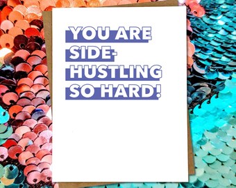May include: A white greeting card with the text "YOU ARE SIDE-HUSTLING SO HARD!" in blue lettering. The card is set against a background of orange and teal sequins.