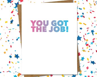 May include: A white greeting card with the text "YOU GOT THE JOB!" in a gradient of pink and blue. The card is set against a background of confetti in various colors and shapes, including stars and rectangles. The card is placed on a brown paper backing.