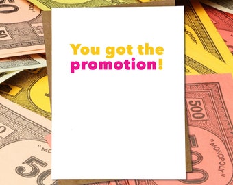 May include: A white greeting card with the text "You got the promotion!" in yellow and pink, set against a backdrop of Monopoly money in various colors. The card has a brown border.
