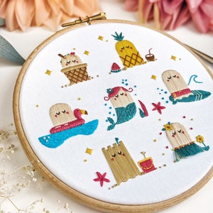 Puede incluir: Arte de bordado en aro con una variedad de personajes de temática veraniega, incluyendo una sirena, una piña, un cono de helado, un castillo de arena y una persona flotando en un flotador de flamenco. Los personajes son todos sonrientes y tienen ojos saltones. El aro está hecho de madera y tiene un fondo de tela blanco.