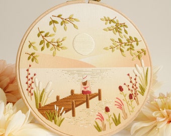 May include: Embroidered hoop art featuring a person sitting on a wooden dock overlooking a lake. The scene is surrounded by green foliage and a white sun.