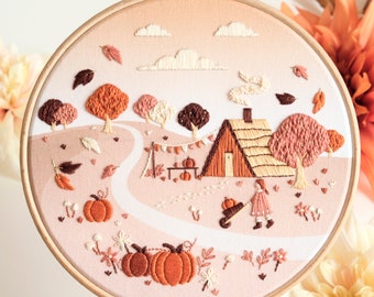 Puede incluir: Arte de bordado en aro que representa una cabaña acogedora en el bosque con un huerto de calabazas y una niña empujando una carretilla. La escena está rodeada de follaje otoñal y flores en tonos naranja, marrón y crema.