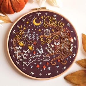 Könnte beinhalten: Runder Stickrahmen mit einer Waldszene mit einem Hirsch, einem Hasen, einer Eule und einem Fuchs. Das Design ist mit weißem, goldenem und orangefarbenem Faden auf einem dunkelburgunderfarbenen Stoff gestickt. Ein Halbmond und Sterne sind ebenfalls enthalten.