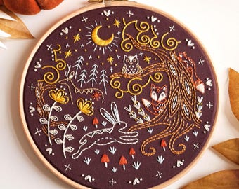 May include: A round embroidery hoop featuring a forest scene with a deer, rabbit, owl, and fox. The design is stitched in white, gold, and orange thread on a dark burgundy fabric. A crescent moon and stars are also included.