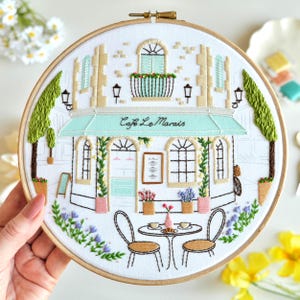 Op de afbeelding: Geborduurde hoepelkunst met een caféscène. Het ontwerp toont een café met een turquoise luifel, de tekst "Cafe Le Marais" en een tafel met twee stoelen. Het kleurenpalet omvat wit, beige, groen, roze en blauw.