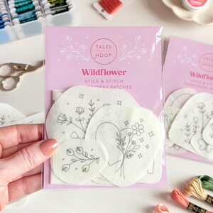 Könnte beinhalten: Eine durchsichtige Kunststoffverpackung mit weißen Stick-and-Stitch-Stickaufnähern aus Stoff mit Wildblumenmotiven. Die rosa Kopfkarte trägt den Text Tales From The Hoop Wildflower Stick & Stitch Embroidery Patches in weißer Schrift.